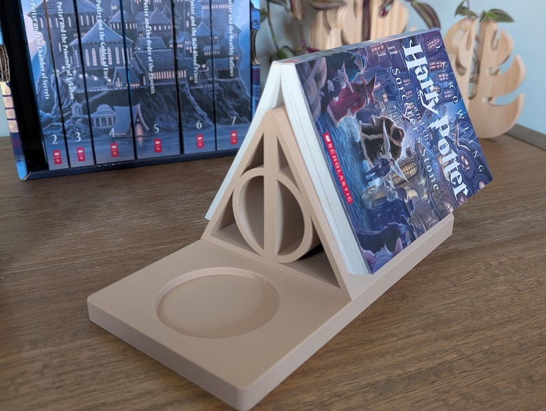 K&ouml;nnte beinhalten: Ein beigefarbener, 3D-gedruckter Buchst&uuml;tze in Form des Harry-Potter-Symbols, den Heiligt&uuml;mern des Todes, mit einem Buch mit dem Titel "Harry Potter und der Stein der Weisen" darauf.