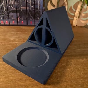 K&ouml;nnte beinhalten: Ein dunkelblauer, 3D-gedruckter Harry-Potter-Telefonst&auml;nder mit einem Symbol der Heiligt&uuml;mer des Todes und einer runden Vertiefung f&uuml;r ein Telefon.