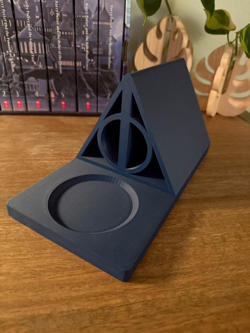 K&ouml;nnte beinhalten: Ein dunkelblauer, 3D-gedruckter Harry-Potter-Telefonst&auml;nder mit einem Symbol der Heiligt&uuml;mer des Todes und einer runden Vertiefung f&uuml;r ein Telefon.