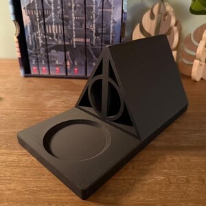 K&ouml;nnte beinhalten: Ein schwarzer, 3D-gedruckter Harry-Potter-Telefonst&auml;nder mit einem Symbol der Heiligt&uuml;mer des Todes und einer runden Vertiefung f&uuml;r ein Telefon.