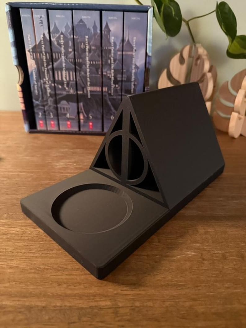 K&ouml;nnte beinhalten: Ein schwarzer, 3D-gedruckter Harry-Potter-Telefonst&auml;nder mit einem Symbol der Heiligt&uuml;mer des Todes und einer runden Vertiefung f&uuml;r ein Telefon.