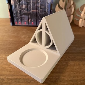 K&ouml;nnte beinhalten: Ein wei&szlig;er Keramik-Telefonst&auml;nder in Form des Symbols der Heiligt&uuml;mer des Todes aus der Harry-Potter-Reihe. Der St&auml;nder hat eine kreisf&ouml;rmige Vertiefung zum Halten eines Telefons.