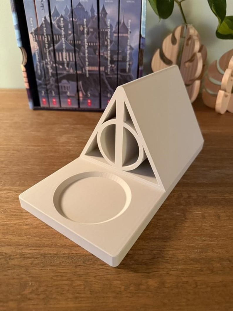 K&ouml;nnte beinhalten: Ein wei&szlig;er Keramik-Telefonst&auml;nder in Form des Symbols der Heiligt&uuml;mer des Todes aus der Harry-Potter-Reihe. Der St&auml;nder hat eine kreisf&ouml;rmige Vertiefung zum Halten eines Telefons.
