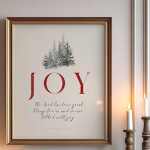 Könnte beinhalten: Ein gerahmter Druck mit weißem Hintergrund und einer roten Aufschrift, die "JOY" lautet. Der Druck zeigt eine Aquarell-Illustration eines schneebedeckten Waldes mit einem Bibelzitat unter dem Bild. Der Druck wird auf einem weißen Kaminsims mit einem grünen Kranz und roten und weißen Ornamenten angezeigt.