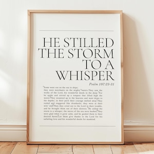 Pode incluir: Uma impressão emoldurada com o texto "HE STILLED THE STORM TO A WHISPER" em uma fonte elegante. A impressão inclui um versículo bíblico, Salmo 107:23-31, e é exibida em um piso de madeira.