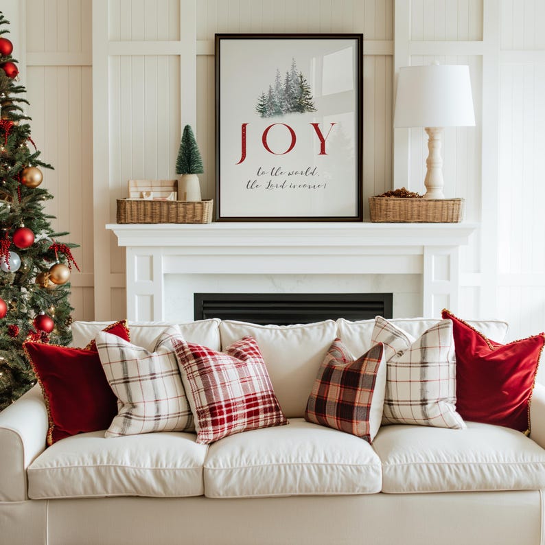 K&ouml;nnte beinhalten: Ein gem&uuml;tliches Wohnzimmer mit einem wei&szlig;en Sofa, das mit roten und karierten Kissen geschm&uuml;ckt ist. Links steht ein Weihnachtsbaum mit Ornamenten. &Uuml;ber dem Kamin h&auml;ngt ein gerahmter Druck mit dem Wort "JOY" und einer Winterlandschaft.