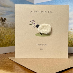 以下が含まれることがあります： 「A Little Note to Say...」と「Thank Ewe」の文字が入ったベージュ色のグリーティングカード。黒い頭と足を持つふわふわの羊が花を持っています。カードは茶色の封筒に入っています。