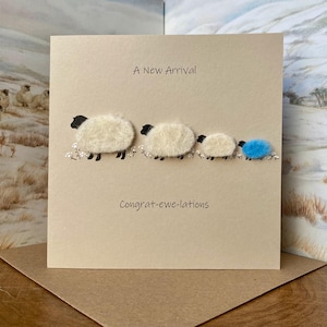 May include: A beige card with the text "A New Arrival" and "Congrat-ewe-lations." It features four sheep, three white and one blue, with black heads and legs, arranged in a line. Small white flowers are painted below.