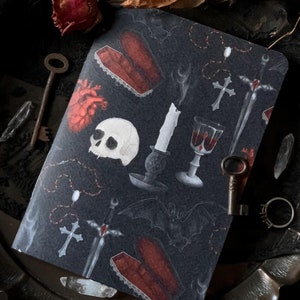 Könnte beinhalten: Ein schwarzes Notizbuch mit einem gotischen Design, das einen Schädel, ein Herz, ein Kreuz, einen Dolch, eine Kerze, einen Kelch und eine Fledermaus zeigt. Das Notizbuch liegt auf einem silbernen Tablett mit einem Schlüssel und einem Ring.