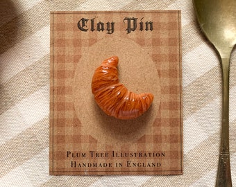 Könnte beinhalten: Eine braune und orangefarbene Tonspange in Form eines Croissants. Die Spange befindet sich auf einer Karte mit dem Text "Clay Pin" und "Plum Tree Illustration Handmade in England".