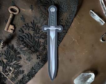 May include: A silver dagger with a black handle, on a dark background with a key and crystals.