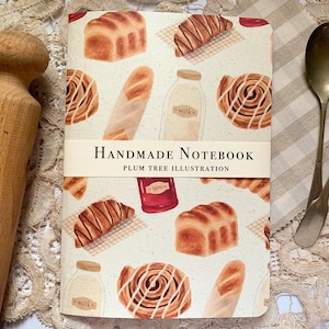 May include: A handmade notebook with a cream-colored cover featuring a repeating pattern of illustrated pastries, bread, and milk bottles. The text "HANDMADE NOTEBOOK" and "PLUM TREE ILLUSTRATION" is printed on a white label across the front of the notebook.