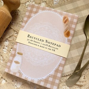 May include: A brown and white checkered notepad with a white lace design and the text "RECYCLED NOTEPAD 50 A6 SHEETS 100% RECYCLED DESIGNED & MADE IN THE UK".