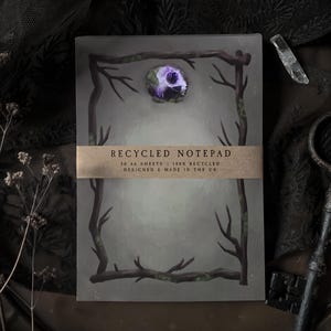 May include: A notepad with a decorative frame of branches and a purple floral accent. The notepad has a brown band with the text "RECYCLED NOTEPAD". The image also includes a magnifying glass and a crystal.