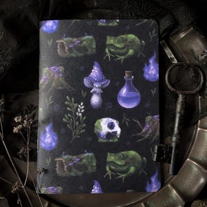 May include: A black notebook featuring a pattern of mushrooms, frogs, and potion bottles. The design includes purple flames, tree stumps, and a skull. The notebook is displayed on a decorative tray with a vintage key.