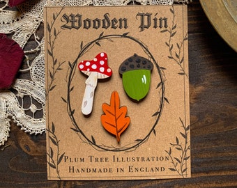 May include: A set of three wooden pins featuring a red and white mushroom, a green acorn, and an orange leaf. The pins are displayed on a brown card with the text "Wooden Pin" and "Plum Tree Illustration Handmade in England".