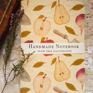 Può includere: Un taccuino fatto a mano con una copertina color crema che presenta un motivo ripetitivo di pere, mele, foglie e attrezzi da giardinaggio. Il testo "HANDMADE NOTEBOOK" e "PLUM TREE ILLUSTRATION" è stampato su un'etichetta bianca sulla parte anteriore del taccuino.
