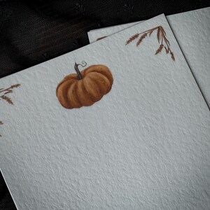 May include: A blank card with a brown pumpkin and brown wheat stalks in the corners. The card is on a textured, light gray surface.
