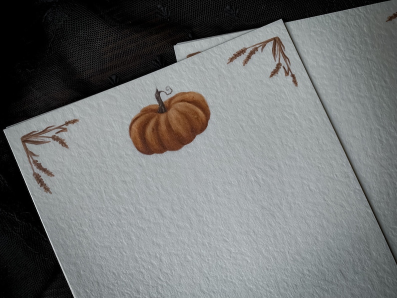 May include: A blank card with a brown pumpkin and brown wheat stalks in the corners. The card is on a textured, light gray surface.