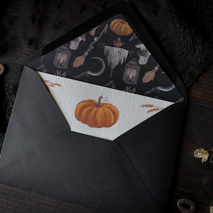 May include: A black envelope with a white card inside. The card has a drawing of a pumpkin on it. The envelope is decorated with a black and white pattern of pumpkins, lanterns, and other Halloween-themed items.