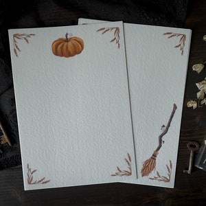 May include: Two blank white stationery sheets with brown borders and fall-themed illustrations. The left sheet features a small orange pumpkin, while the right sheet has a brown broom with a brown handle.