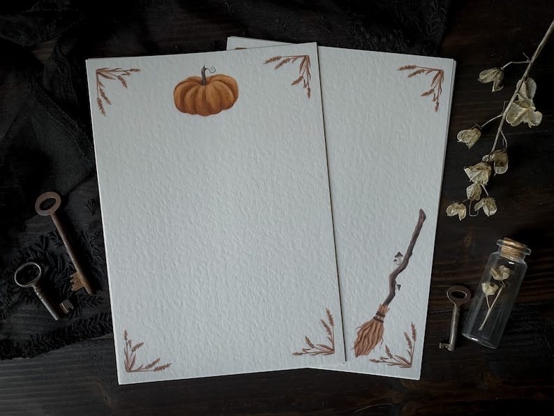 May include: Two blank white stationery sheets with brown borders and fall-themed illustrations. The left sheet features a small orange pumpkin, while the right sheet has a brown broom with a brown handle.