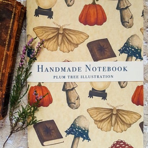 Op de afbeelding: Een handgemaakt notitieboek met een crèmekleurige kaft met een herhalend patroon van motten, pompoenen, paddenstoelen en kristallen bollen. De tekst "HANDMADE NOTEBOOK" en "PLUM TREE ILLUSTRATION" staat op een witte band.