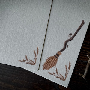 May include: A blank card with a brown and tan illustration of a broom with a few small mushrooms on the handle. The card is on a dark wood surface with a key and a glass jar with dried flowers.
