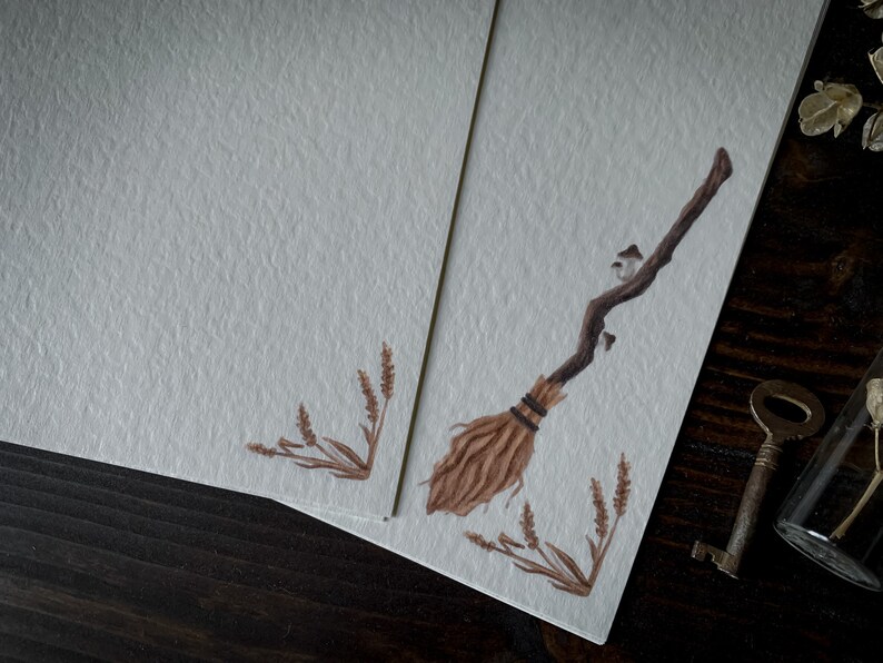 May include: A blank card with a brown and tan illustration of a broom with a few small mushrooms on the handle. The card is on a dark wood surface with a key and a glass jar with dried flowers.