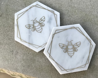May include: Two white marble hexagon-shaped coasters with gold bee designs. The bees are outlined in gold and have a geometric pattern.