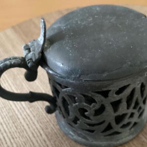 Könnte beinhalten: Ein Vintage-Behälter aus Zinn mit Deckel und Griff. Der Behälter hat ein durchbrochenes Design an den Seiten und einen dekorativen Griff. Der Deckel hat einen kleinen Griff. Das Zinn hat ein dunkles, gealtertes Aussehen.
