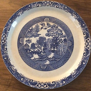 Puede incluir: Un plato redondo de cerámica blanca y azul con un detallado patrón Willow. El plato presenta una escena central con una pagoda, árboles y un puente, rodeada por un borde decorativo. El borde del plato tiene un diseño floral azul.