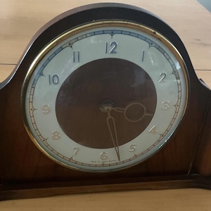 May include: A vintage wooden mantel clock with a light-colored face and dark brown center. The clock face has black numerals and gold-colored trim. The clock's wooden frame is a dark brown color.