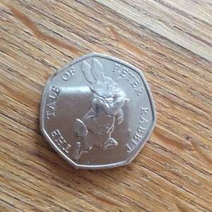 Puede incluir: Una moneda plateada de cincuenta peniques con una ilustración de Peter Rabbit. La moneda tiene la inscripción "THE TALE OF PETER RABBIT". La moneda está sobre una superficie de madera.