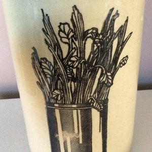 Może przedstawiać: Kremowy ceramiczny kubek z czarną ilustracją łodyg selera w pojemniku. Słowo "CELERY" jest wydrukowane na dole na czarno. Kubek ma lekko zwężający się kształt.