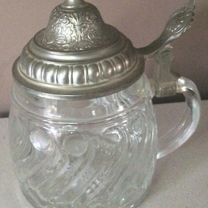 Op de afbeelding: Een helder glazen bierpul met een zilveren deksel en handvat. De pul heeft een decoratief patroon van spiralen en cirkels.