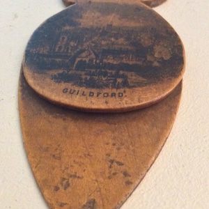 Puede incluir: Un recuerdo de madera vintage con una forma única, que presenta una ilustración detallada en blanco y negro de una escena urbana. La palabra "GUILDFORD" está impresa en la parte delantera. El artículo tiene un aspecto desgastado y envejecido.