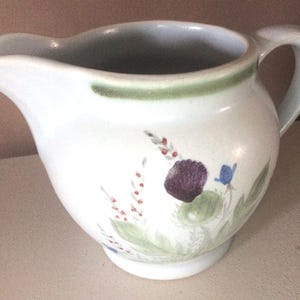Może przedstawiać: Biały dzbanek do śmietanki z ceramiczną rączką, zielonym brzegiem i kwiatowym wzorem z fioletowymi, niebieskimi i zielonymi kwiatami.
