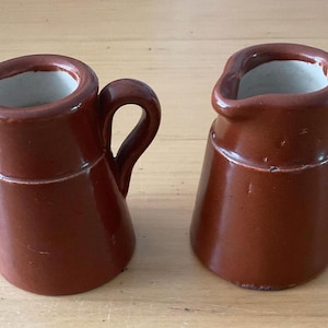 May include: Two small, brown ceramic pitchers with handles. The pitchers have a classic design with a spout for pouring and a white interior. They are likely used for serving cream or other liquids.