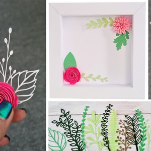 May include: A white frame with a pink paper flower and green leaves inside. There are additional pink paper flowers and green leaves on a white surface.