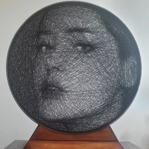 Può includere: Un'opera d'arte a filo nero rotonda raffigurante un volto, montata su una base di legno. Il volto è creato con filo nero e la base in legno è di colore marrone caldo. Un oggetto di arredamento unico.