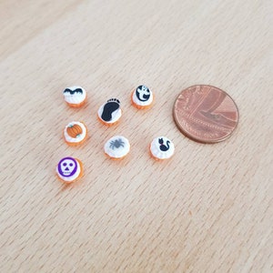 May include: A collection of miniature Halloween-themed decorations. The decorations include a pumpkin, a ghost, a bat, a spider, a skull, a footprint, and a black cat. The decorations are orange and white, and are next to a penny for size comparison.