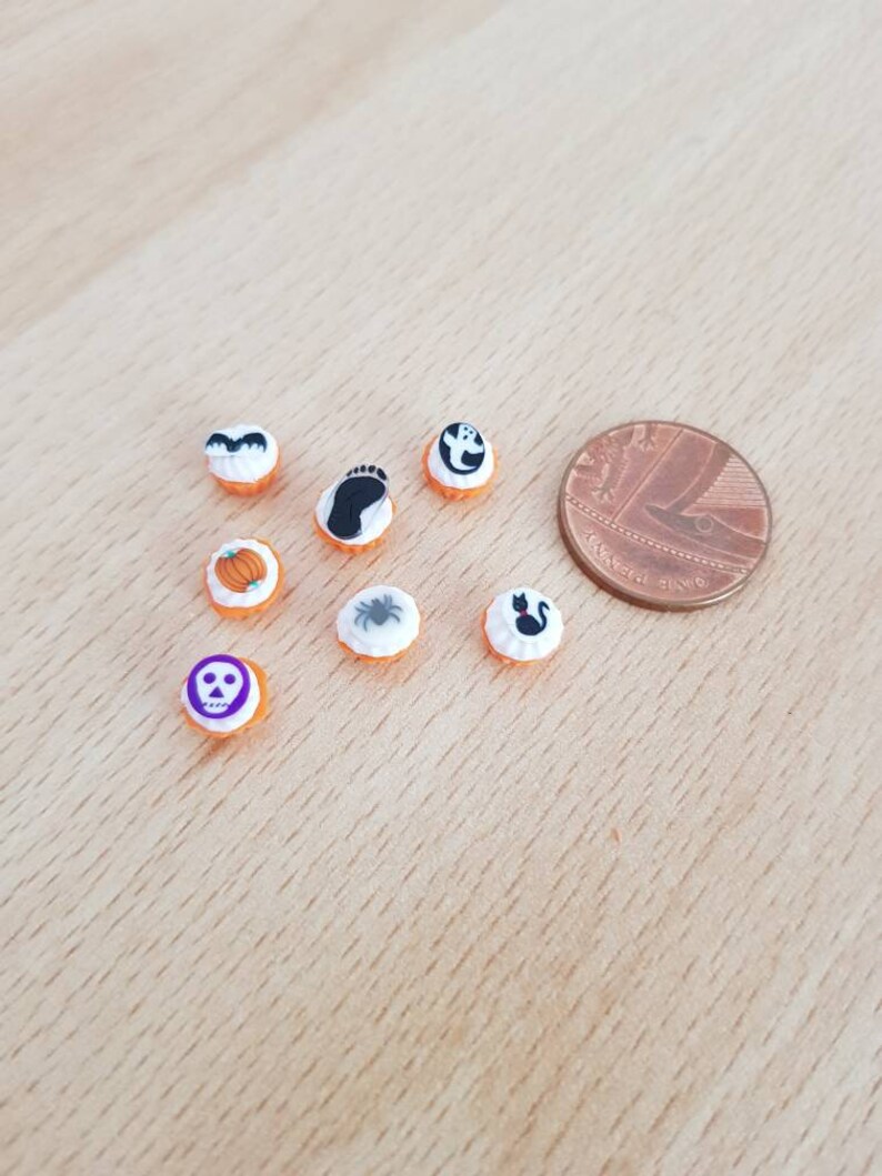 May include: A collection of miniature Halloween-themed decorations. The decorations include a pumpkin, a ghost, a bat, a spider, a skull, a footprint, and a black cat. The decorations are orange and white, and are next to a penny for size comparison.