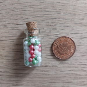 May include: A small glass bottle with a cork stopper filled with white, green, and red pearl sprinkles. The bottle is next to a British 1 pence coin.
