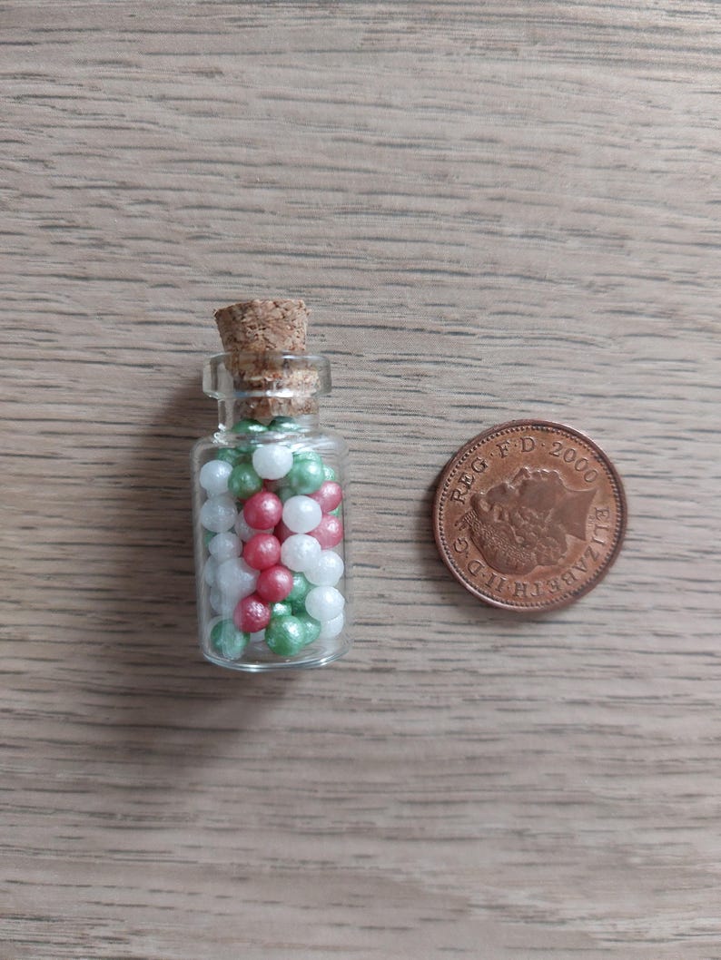 May include: A small glass bottle with a cork stopper filled with white, green, and red pearl sprinkles. The bottle is next to a British 1 pence coin.
