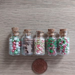 May include: Five small glass jars with cork lids filled with various Christmas sprinkles and cookie shapes. The jars are lined up on a wood surface.