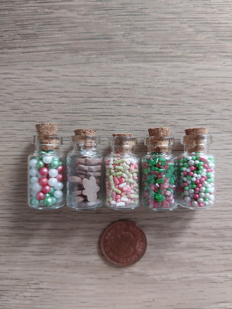 May include: Five small glass jars with cork lids filled with various Christmas sprinkles and cookie shapes. The jars are lined up on a wood surface.