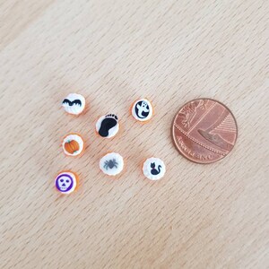May include: A collection of small, round Halloween-themed embellishments. The designs include a bat, footprint, ghost, pumpkin, spider, skull, and a black cat. A penny is included for size reference. The embellishments are white with orange borders.