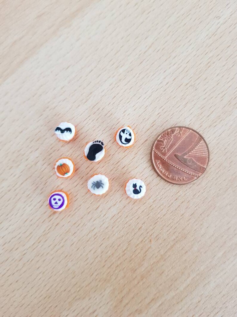 May include: A collection of small, round Halloween-themed embellishments. The designs include a bat, footprint, ghost, pumpkin, spider, skull, and a black cat. A penny is included for size reference. The embellishments are white with orange borders.