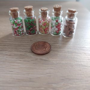 May include: Five small glass jars with cork lids filled with various colorful sprinkles and cookie shapes. The jars are lined up on a wooden surface with a copper coin in front of them.